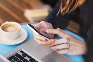 A woman is sitting in front of her laptop holding her mobile phone.  Next to her is a coffee cup.  She is looking at the new P&A retirement plan web portal.