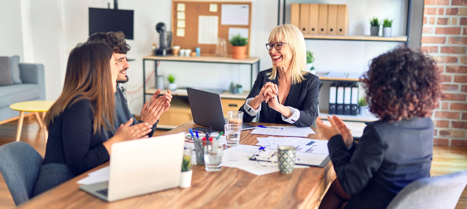 A group of colleagues are sitting around a rectangular conference table with papers spread out and a laptop. The co-workers are engaged in a lively discussion and are smiling.